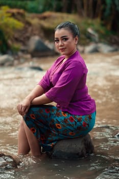 Beautiful Asian woman sitting on the rock near the river with a smiling face while wearing a purple dress inside the village