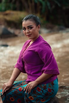 Beautiful Asian woman sitting on the rock near the river with a smiling face while wearing a purple dress inside the village