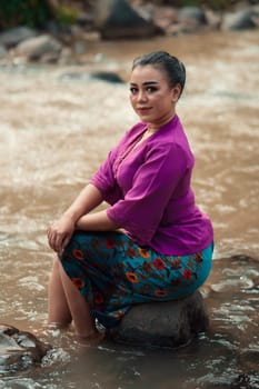Beautiful Asian woman sitting on the rock near the river with a smiling face while wearing a purple dress inside the village