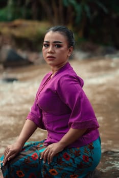 Beautiful Asian woman sitting on the rock near the river with a smiling face while wearing a purple dress inside the village