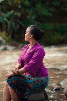 Indonesian woman with purple clothes and makeup on her face sitting near the river with brown color water while visiting the river for vacation