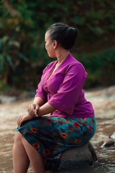 Indonesian woman with purple clothes and makeup on her face sitting near the river with brown color water while visiting the river for vacation
