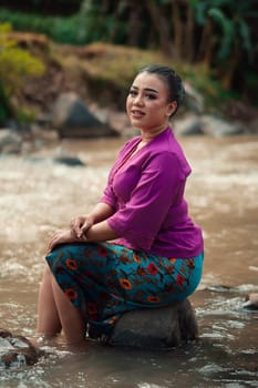 Indonesian woman with purple clothes and makeup on her face sitting near the river with brown color water while visiting the river for vacation