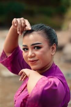 Beautiful Javanese woman with makeup and purple dress posing with her hand while sitting on the rock near the river inside the village