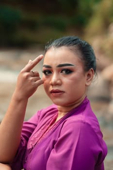 Beautiful Javanese woman with makeup and purple dress posing with her hand while sitting on the rock near the river inside the village