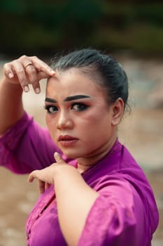 Beautiful Javanese woman with makeup and purple dress posing with her hand while sitting on the rock near the river inside the village