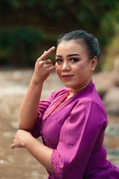 Beautiful Javanese woman with makeup and purple dress posing with her hand while sitting on the rock near the river inside the village