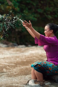 Happy Asian woman playing with the water near the river while sitting on the rock and wearing a purple dress inside the village