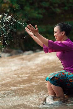 Happy Asian woman playing with the water near the river while sitting on the rock and wearing a purple dress inside the village