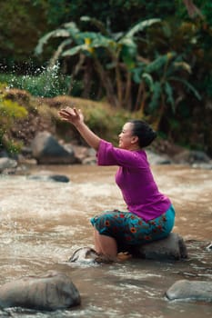 Happy Asian woman playing with the water near the river while sitting on the rock and wearing a purple dress inside the village