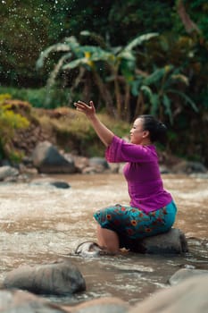 Indonesian woman sitting on a small rock and playing with the water while her dress got wet from the river near the rice field