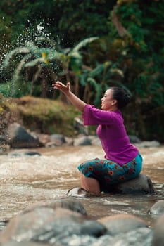 Indonesian woman sitting on a small rock and playing with the water while her dress got wet from the river near the rice field