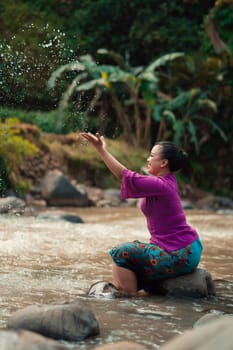 Indonesian woman sitting on a small rock and playing with the water while her dress got wet from the river near the rice field