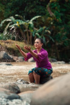 Asian woman playing with dirty water from a dirty river while wearing a purple dress and green skirt near the river