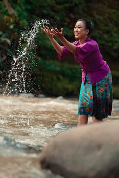 A beautiful Asian woman washing her hand and playing with the water while standing near the river in a traditional purple dress and green skirt in the village