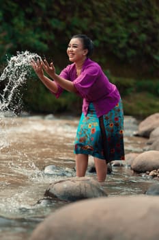 A beautiful Asian woman washing her hand and playing with the water while standing near the river in a traditional purple dress and green skirt in the village