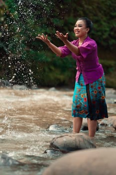 A beautiful Asian woman washing her hand and playing with the water while standing near the river in a traditional purple dress and green skirt in the village