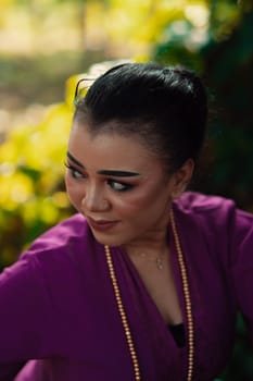 The face of a Javanese woman with makeup and a traditional purple dress called kebaya while dancing and posing with jewelry on her neck inside the jungle