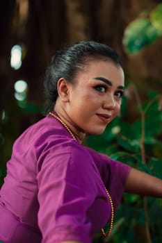 The face of a Javanese woman with makeup and a traditional purple dress called kebaya while dancing and posing with jewelry on her neck inside the jungle
