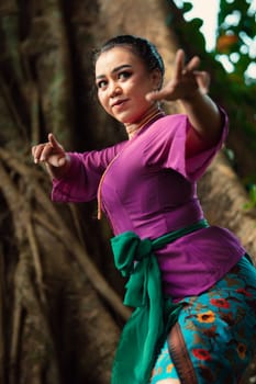 Sexy Asian woman dancing pose while wearing a beautiful purple dress and green belt on her body in front of the big brown tree inside the forest