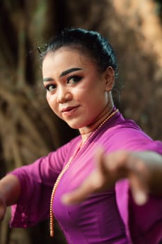 The face of a Javanese woman with makeup and a traditional purple dress called kebaya while dancing and posing with jewelry on her neck inside the jungle