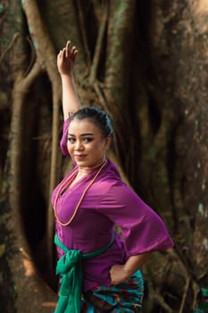 An Indonesian woman bravely poses while raising her hands in a purple dress while standing in the forest