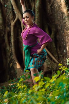Asian woman takes a dancing pose in the jungle while wearing a purple dress and a golden necklace with a beautiful smile on her face