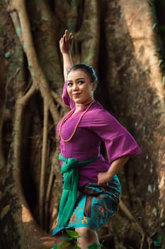 Asian woman takes a dancing pose in the jungle while wearing a purple dress and a golden necklace with a beautiful smile on her face