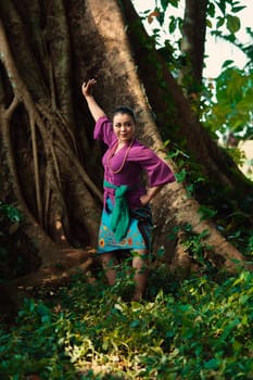 An Indonesian woman bravely poses while raising her hands in a purple dress while standing in the forest