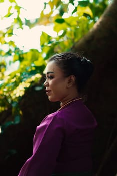 An Asian woman beautifully sitting on the big tree in a purple dress while visiting the jungle in the daylight