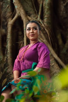 An Asian woman beautifully sitting on the big tree in a purple dress while visiting the jungle in the daylight