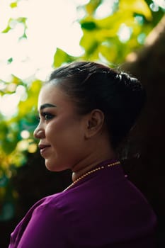 Indonesian woman smiling in a traditional purple dress while wearing makeup and golden jewelry in front of the big tree inside the forest