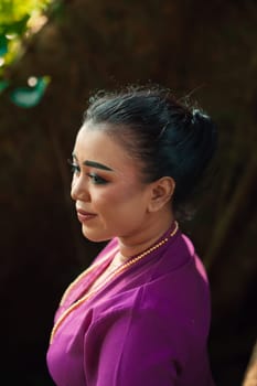 Indonesian woman smiling in a traditional purple dress while wearing makeup and golden jewelry in front of the big tree inside the forest
