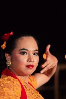 Balinese woman with red lips and makeup wearing a yellow dress and red scarf on a dark night inside the village
