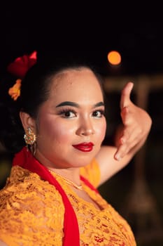 Balinese woman with red lips and makeup wearing a yellow dress and red scarf on a dark night inside the village