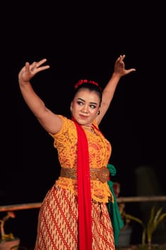 Balinese woman in traditional orange dress dancing with a red scarf while performing dancing in the night