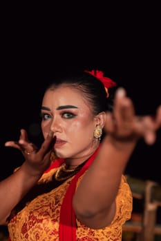 the fierce face of a Balinese woman with makeup in a traditional orange dress and red scarf while dancing at the festival in the night