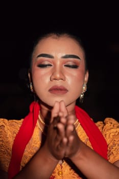 Asian woman praying with both her hands while wearing makeup and an orange dress with a red scarf on her body in the dark night