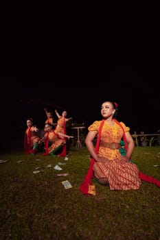 A Group of Indonesian dancers performing on the stage with a red scarf and traditional orange dress inside the festival during the night