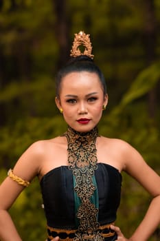 Balinese woman in makeup and gold jewelry on her body with a black costume standing in front of the jungle