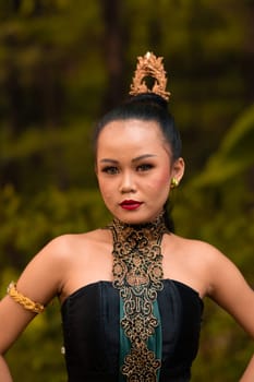Balinese woman in makeup and gold jewelry on her body with a black costume standing in front of the jungle