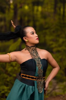 Gorgeous Asian woman in makeup with black hair and red lipstick wearing golden jewelry during the dance performance in front of the jungle