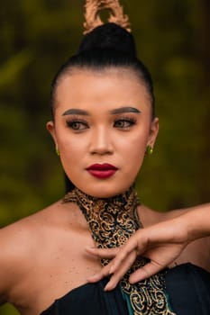 Portrait of an Asian woman in makeup and red lips while wearing a golden necklace and golden accessories on her black hair while standing in front of the jungle