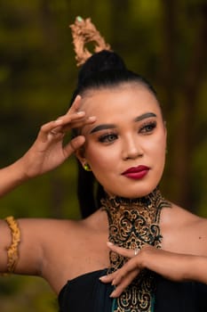 Portrait of an Asian woman in makeup and red lips while wearing a golden necklace and golden accessories on her black hair while standing in front of the jungle