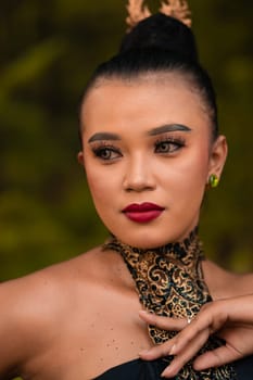 Beautiful eyes of an Asian woman in makeup and red lipstick while wearing gold jewelry and black hair while standing in front of the jungle
