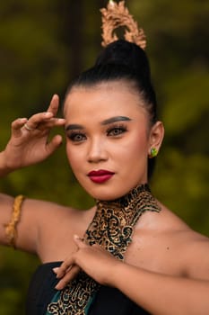 Portrait of an Asian woman in makeup and red lips while wearing a golden necklace and golden accessories on her black hair while standing in front of the jungle