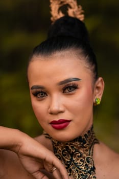 Beautiful eyes of an Asian woman in makeup and red lipstick while wearing gold jewelry and black hair while standing in front of the jungle