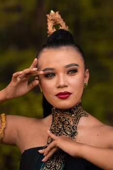 Portrait of an Asian woman in makeup and red lips while wearing a golden necklace and golden accessories on her black hair while standing in front of the jungle