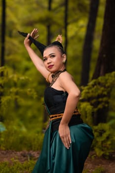 Asian woman holding her black hair in a green costume while wearing makeup and posing in front of the forest during the dance festival inside the village