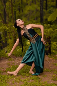Beautiful Asian woman in a green costume with long black hair dances in the woods during the daylight